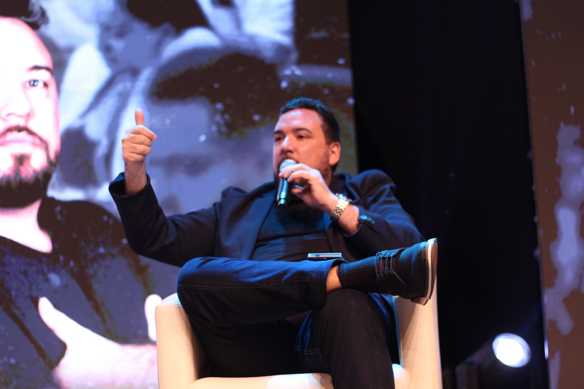 Romulo Vieira sentado em painel no palco, falando ao microfone