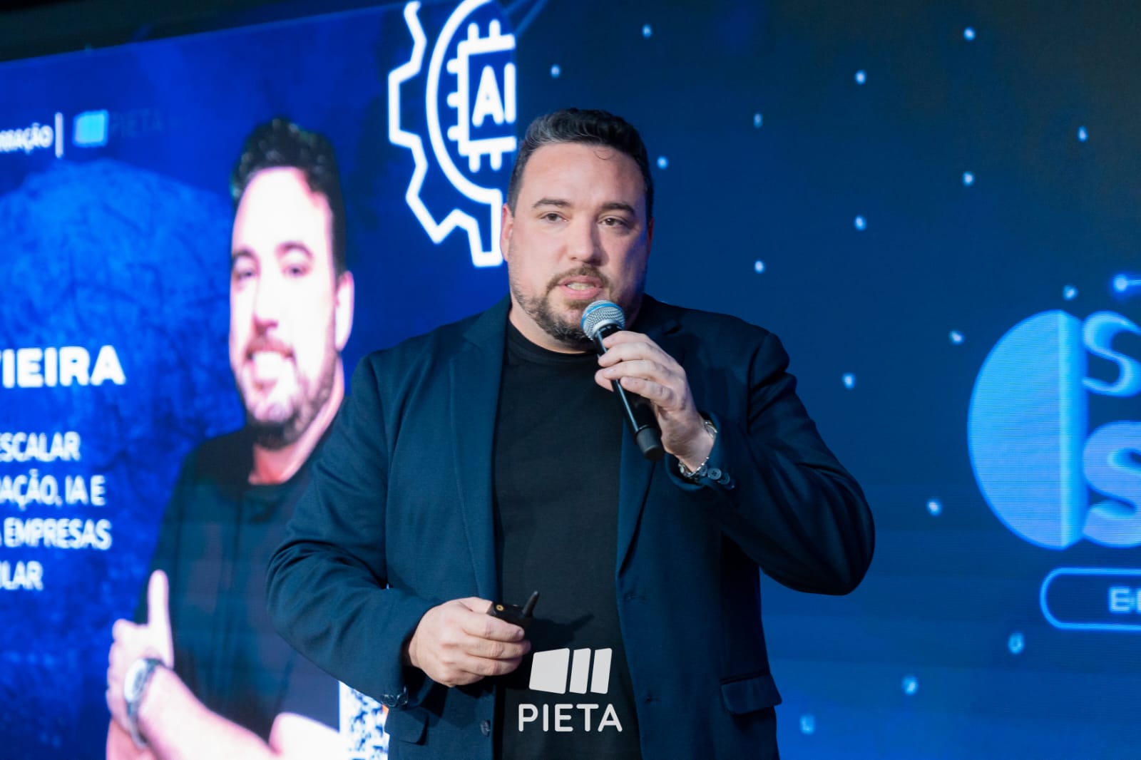 Romulo Vieira ao microfone diante de um telão durante apresentação